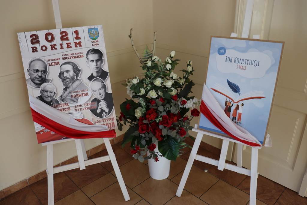 Na zdjęciu plakaty informujące o święcie Konstytucji 3 Maja. Plakaty udekorowane flagą Polski. Pomiędzy plakatami stoi wazon z różami w barwach narodowych.