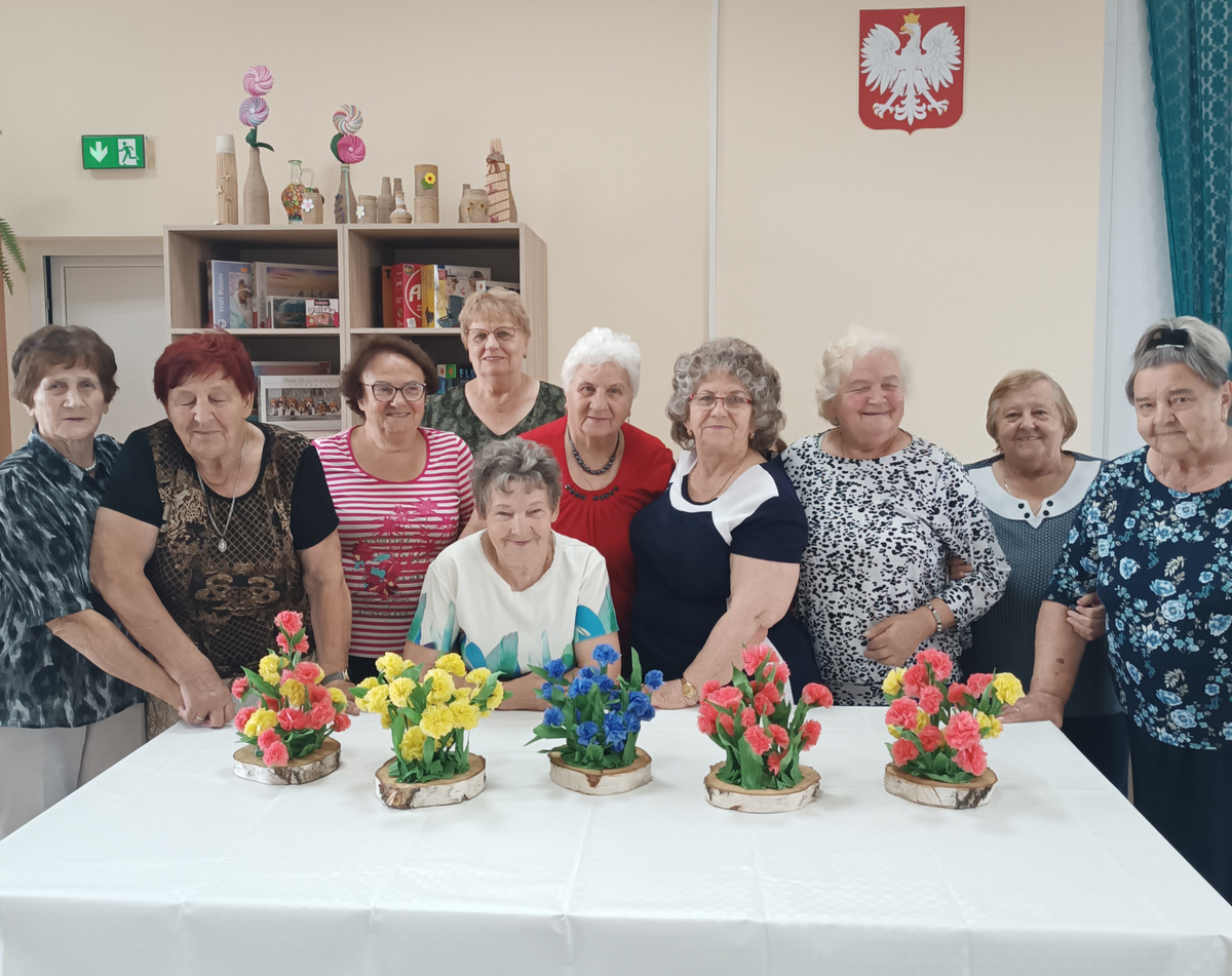 Seniorzy z wykonywanymi kwiatami z folii na plastrze brzozy.