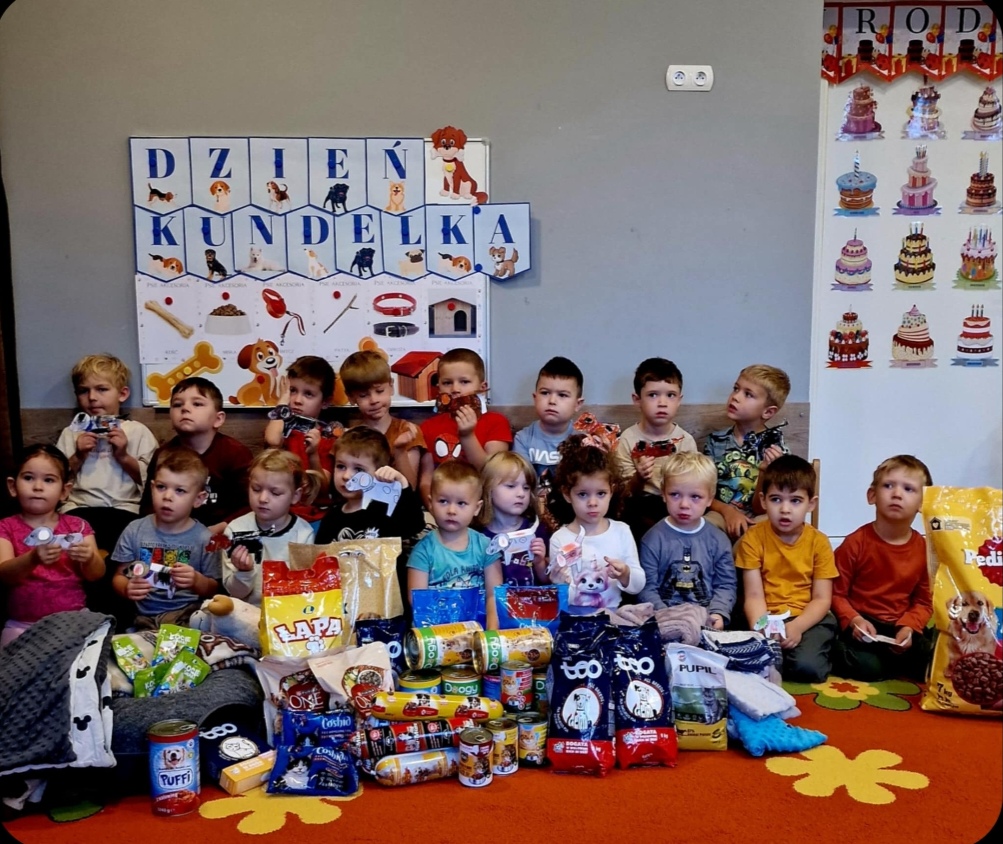Dzieci z grupy Biedronki i Motylki przy tablicy tematycznej, z wcześniej przyniesioną karmą dla zwierząt.
