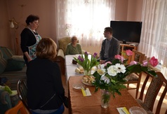 Uczestnicy spotkania na czele z solenizantką siedzą przy stole, na których stoją urodzinowe kwiaty.